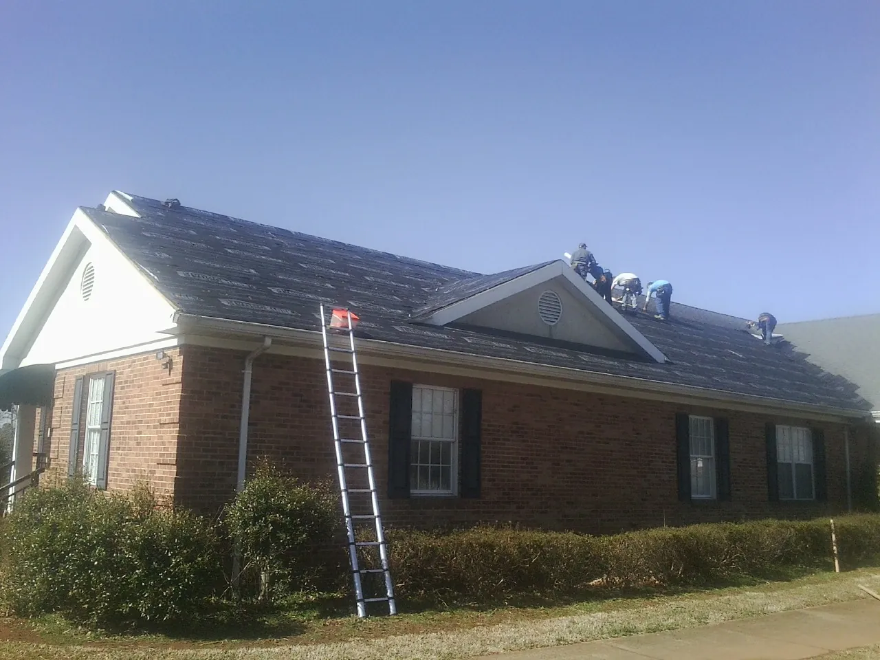 Crew tearing shingles off a brick ranch roof with a ladder set — Emergency Roof Repair work by our crew in Fitchburg