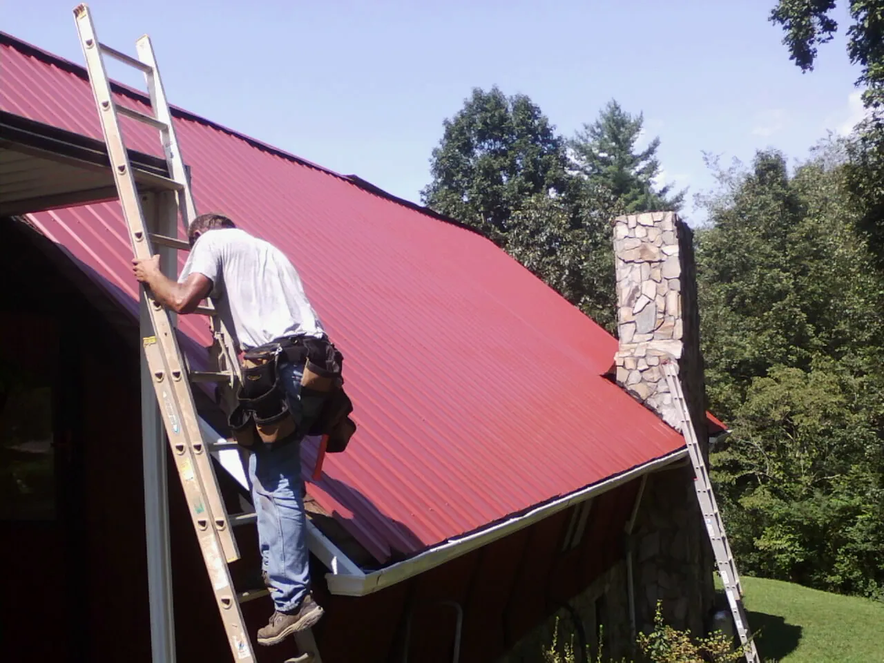 Roofer on a ladder beside a red metal roof and stone chimney — Soffit & Fascia Repair by Roofing Brothers in Fitchburg